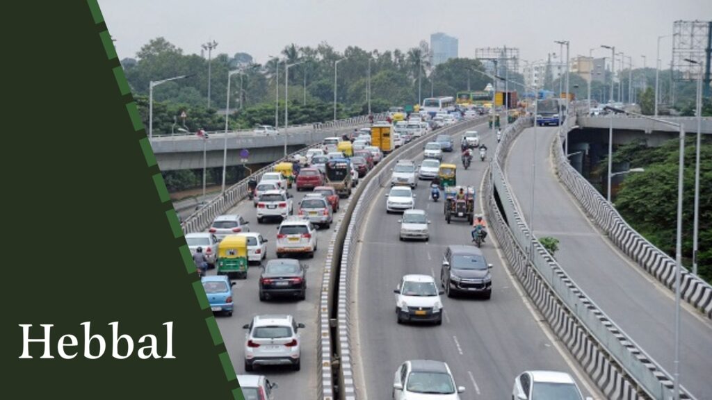 A busy road with multiple lanes of traffic, including cars and auto-rickshaws, on an elevated expressway in Hebbal, flanked by trees and buildings in the background, highlights the bustling activity and rapid development driven by house construction companies in Bangalore.